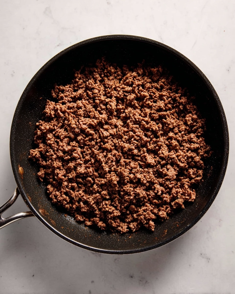 A black pan filled with cooked ground beef covering most of the pan’s flat surface. The beef has a crumbly texture and a dark brown color with small hints of lighter brown areas, evenly spread out in one layer. The black pan has two handles, one on each side, and sits on a white marbled surface. The lighting highlights the texture and moisture slightly shining on the beef. photo taken with an iphone --ar 4:5 --v 7