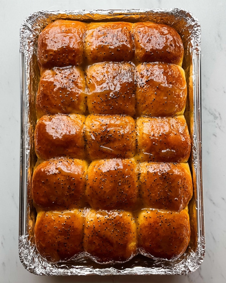 The image shows a rectangular glass pan lined with crinkled aluminum foil, filled with 15 small soft bread rolls arranged in three rows of five. Each roll is golden brown and has a shiny, thick glaze on top, dotted with specks of black seasoning. The rolls are tightly packed, with the glaze creating a glossy, slightly sticky surface that enhances their rich brown color. The pan sits on a white marbled surface. photo taken with an iphone --ar 4:5 --v 7
