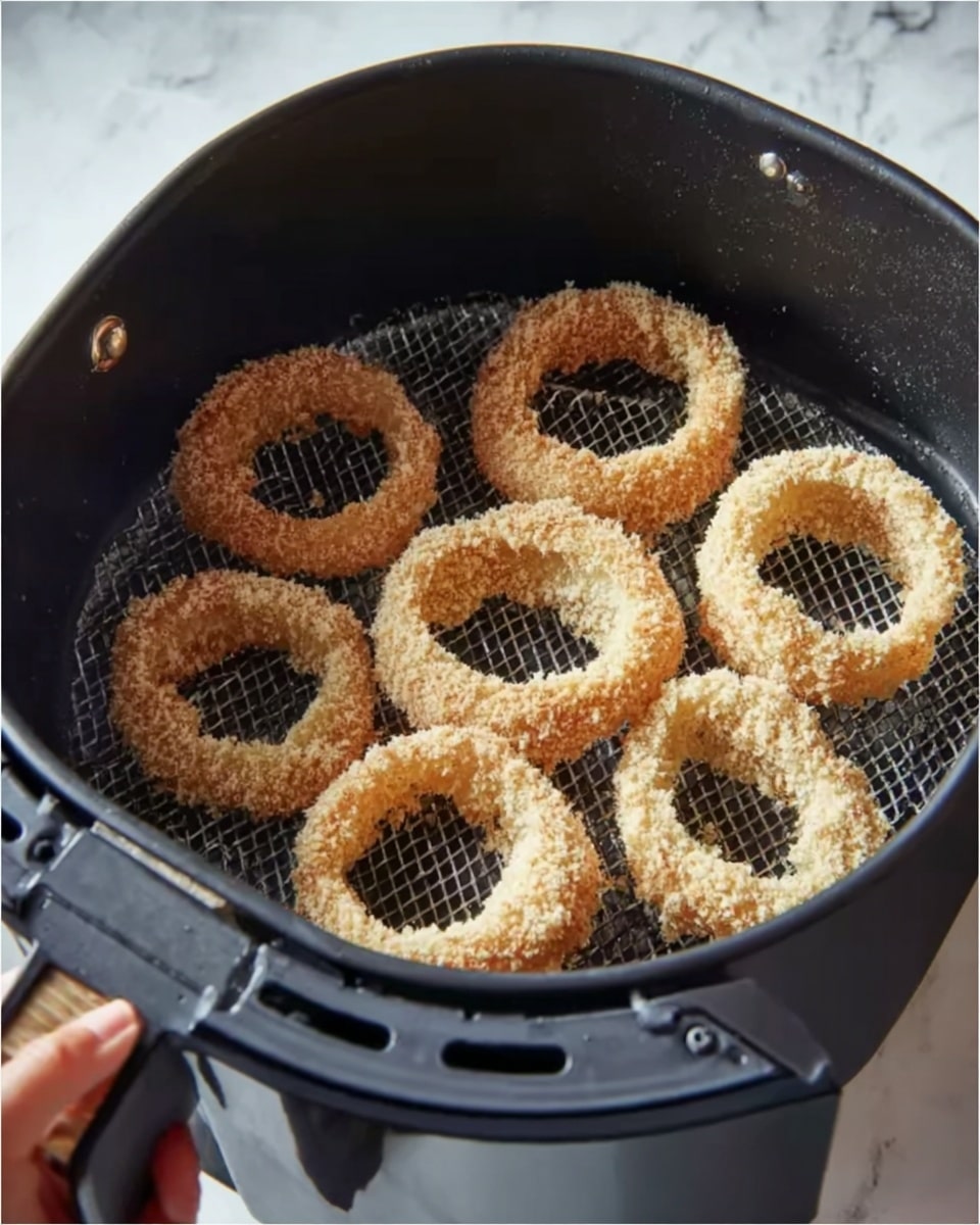 The image shows a black air fryer basket filled with seven onion rings. Each onion ring is coated in a crispy, light golden breading with a textured surface that looks crunchy. The rings are arranged to fit closely but not stacked, with some overlapping edges visible. The inside of the basket has a black mesh base and sides that curve upward, containing the rings. A woman's hand is holding the basket by a black handle with a touch of brown on the side. The background and surface are a white marbled texture. Photo taken with an iphone --ar 4:5 --v 7