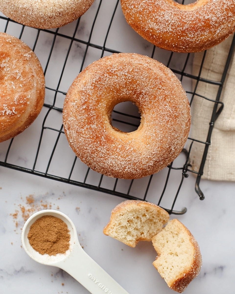 The image shows three light brown doughnuts on a black cooling rack with sugar and cinnamon dusted on top giving a slightly grainy texture. One doughnut is whole and placed near the center, another is partially visible at the top left, and the third doughnut is broken into two pieces at the top right, showing a soft, fluffy inside with a slightly rough texture. Below the cooling rack, there is a white measuring spoon with ground cinnamon resting on a white marbled surface. The colors are warm and light with a clean, fresh look. photo taken with an iphone --ar 4:5 --v 7