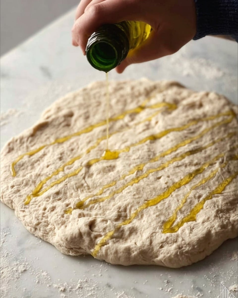 A close-up view of a raw, round dough spread thin on a white marbled surface with a slightly rough texture. A woman's hand is seen squeezing oil from a green bottle, drizzling golden-yellow oil in wide zigzag lines across the dough’s uneven surface. The dough is pale beige with subtle air pockets and small flour patches. Photo taken with an iphone --ar 4:5 --v 7