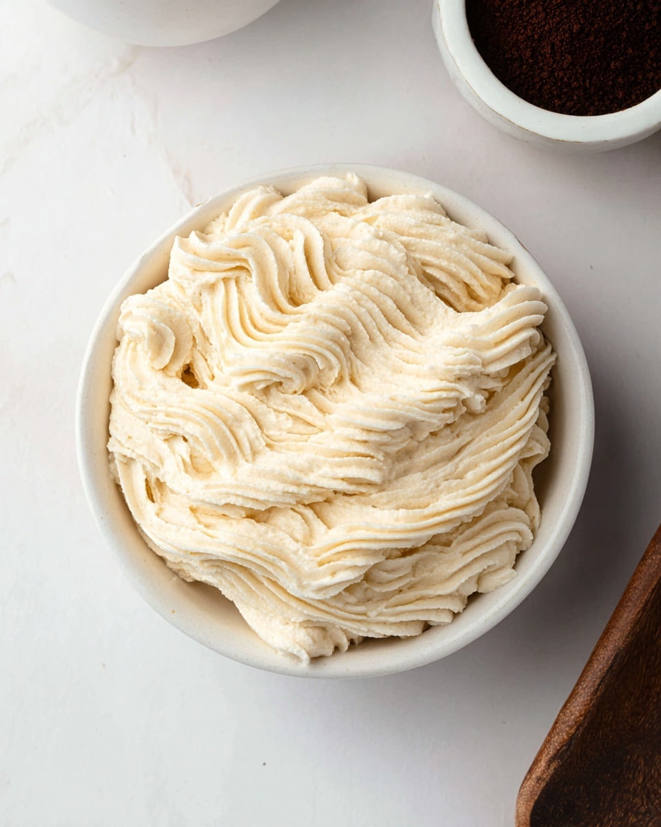 A white round bowl filled with a single layer of light beige cream piped in thick, wavy lines closely packed together, showing a soft and airy texture. The bowl is placed on a white marbled surface, with a portion of another white bowl containing dark brown powder visible in the upper right corner and a wooden object in the upper left corner. The cream has a smooth, slightly ridged appearance from the piping. Photo taken with an iphone --ar 4:5 --v 7