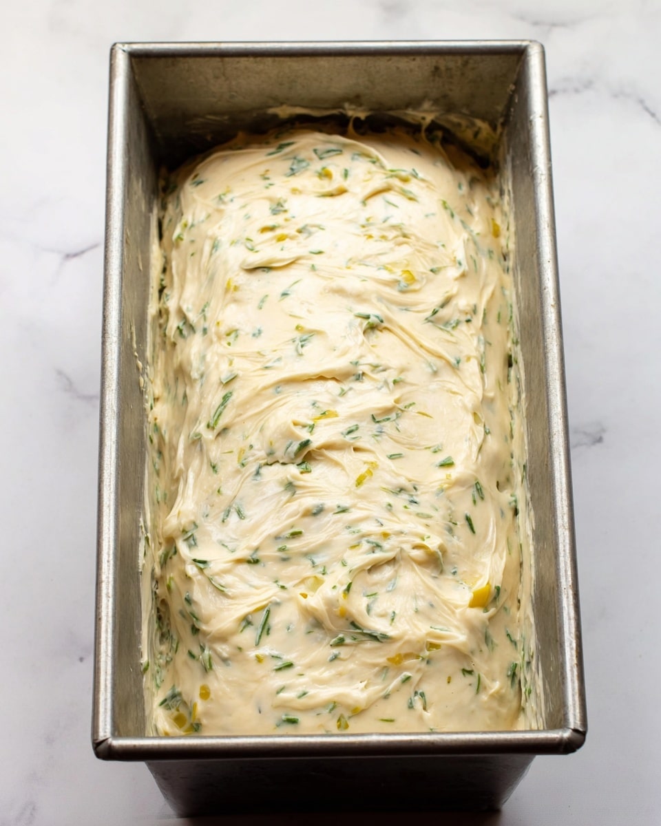 The image shows a rectangular metal baking pan filled with a single large loaf covered evenly in a thick creamy mixture. The creamy layer is light beige with small green and yellow bits mixed throughout, giving it a textured appearance. The loaf is smooth but has some gentle ridges and patches where the creamy mixture is spread with visible brush strokes. The pan rests on a white marbled surface. photo taken with an iphone --ar 4:5 --v 7