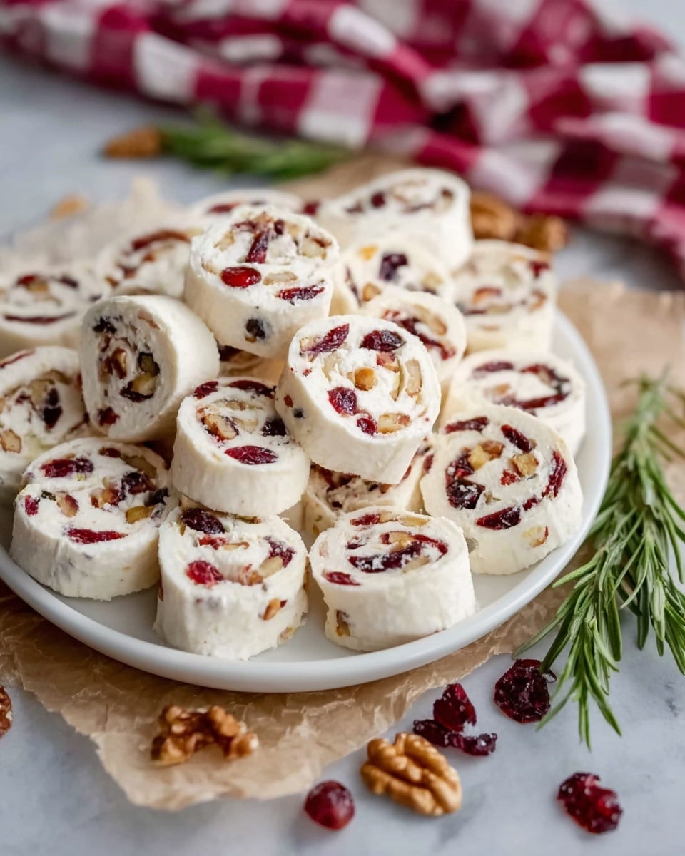 A white round plate is filled with about 20 small pinwheel slices, each with a creamy white outer layer and a filling that has red dried cranberries and some small pieces of nuts mixed inside. The pinwheels are stacked somewhat randomly, showing the spiral pattern of each slice clearly. The plate sits on some crumpled beige parchment paper, surrounded by scattered pieces of walnuts and dried cranberries on a white marbled surface. There is a sprig of green rosemary placed on the right side of the plate. The background includes a red and white checkered cloth that is softly blurred. Photo taken with an iphone --ar 4:5 --v 7