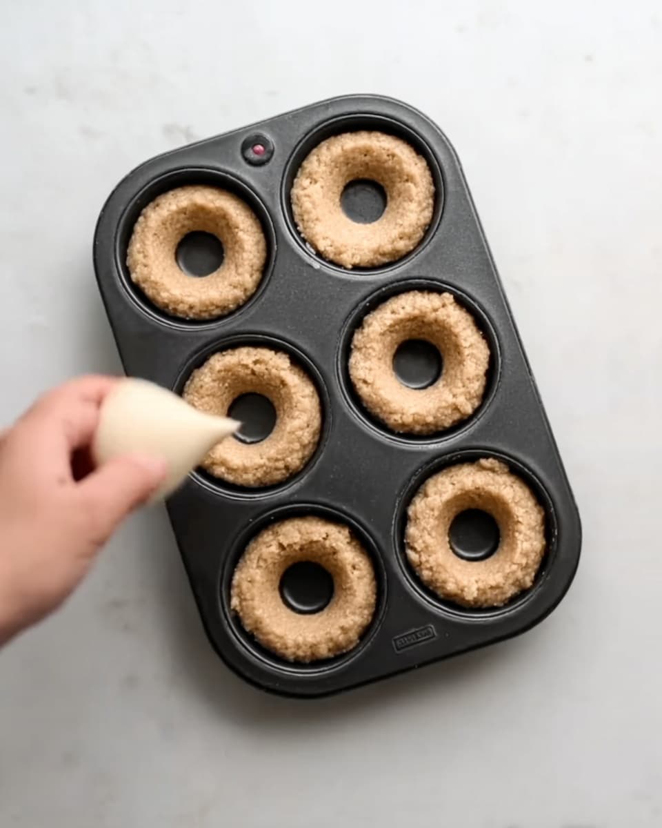 A black metal donut pan with six round cavities sits on a white marbled surface. Five cavities are filled with light brown dough, each with a hole in the middle, showing a slightly rough texture. The sixth cavity is being filled with more dough by a woman's hand holding a piping bag, squeezing the dough into the empty space. The lighting is soft and natural, highlighting the dough's grainy texture and the pan's smooth surface. Photo taken with an iphone --ar 4:5 --v 7