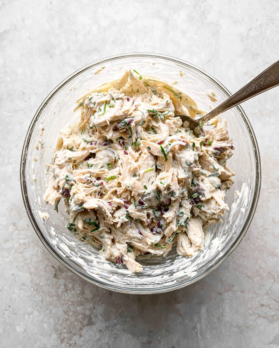 A clear glass bowl sits on a white marbled surface, filled with a creamy mixture that has three main parts: shredded light beige pieces, likely chicken, mixed with small dark purple bits and thin green herb strips. The creamy texture is white with speckles of green and purple, showing the herbs and small vegetable pieces blended throughout. A silver spoon rests inside the bowl, partially covered by the mixture. The image gives a close, top-down view showing the mixed layers clearly, with the bowl’s round edges softly reflecting light. photo taken with an iphone --ar 4:5 --v 7