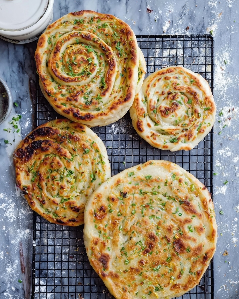 The image shows four round flatbreads with green herbs inside, placed on a black cooling rack. There are two large flatbreads and two smaller ones. Each flatbread has a spiral pattern inside, with layers of light golden dough and scattered green bits. The bigger flatbreads have a few browned spots on the surface, and the smaller ones are more evenly browned, with some crisp edges. The background is a white marbled surface with a dusting of flour. A white dish with a lid is partly visible in the upper left corner. Photo taken with an iphone --ar 4:5 --v 7