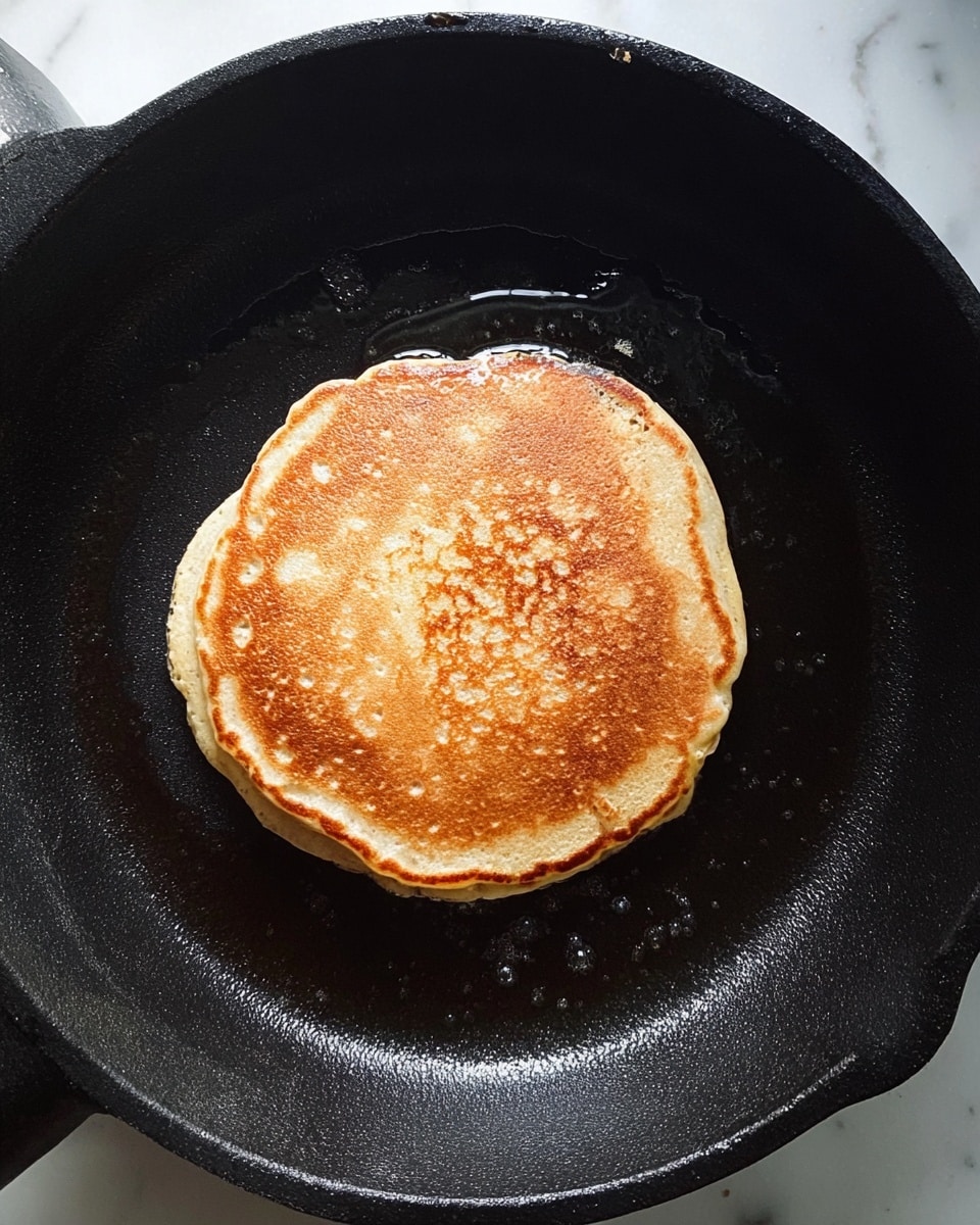 A single pancake is cooking in the center of a black iron pan, showing a light golden-brown surface with small bubbles and a slightly darker round edge that nearly touches the skillet's center. The pancake's texture looks soft with subtle uneven spots, and a bit of light butter or oil is seen sizzling around it. The black pan has a matte finish and slight shine from the cooking surface. The background is a white marbled texture. Photo taken with an iphone --ar 4:5 --v 7