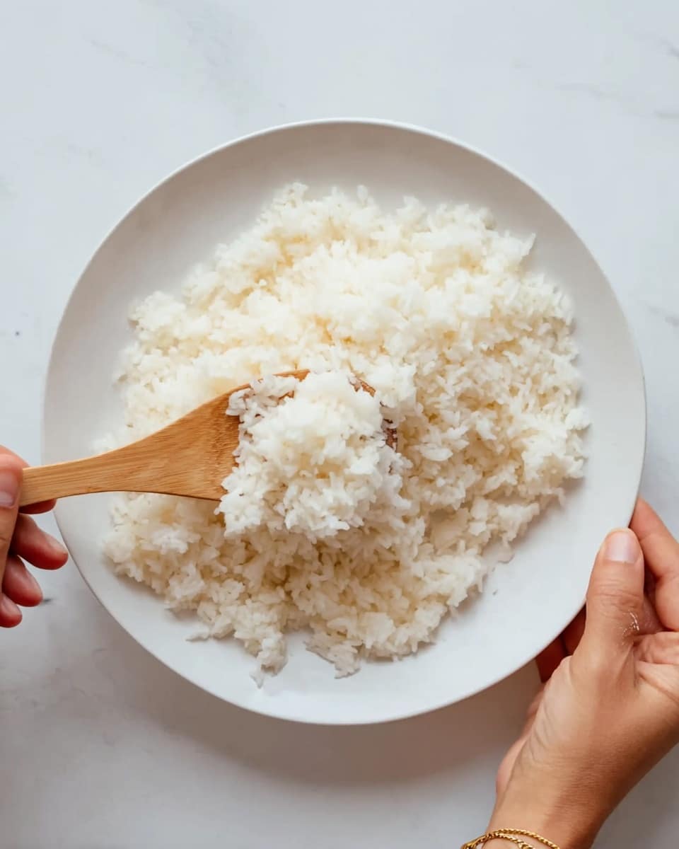 A white round plate sits on a white marbled surface, filled with a single layer of soft white rice grains that look slightly sticky and clumped together. A woman's hand holds a light wooden spatula, positioned near the bottom center of the plate, gently scooping or spreading the rice. Another woman's hand, wearing a thin gold bracelet with a small round charm, is seen from the top right corner, adjusting or holding the edge of the plate. The overall look is clean and simple with a focus on the texture and softness of the rice. photo taken with an iphone --ar 4:5 --v 7