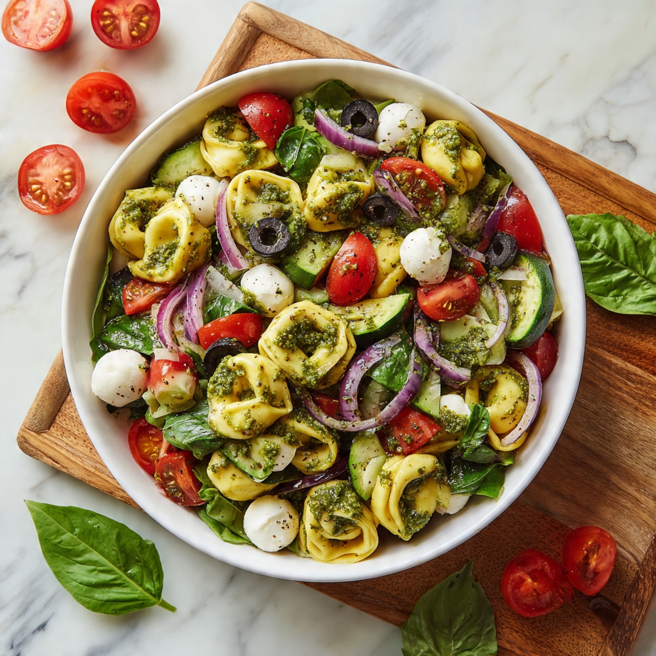 A white bowl filled with a fresh tortellini salad sits on a wooden board over a white marbled surface. The salad has three main layers: the first layer is yellow tortellini pasta with a soft texture, mixed throughout; the second layer consists of halved red cherry tomatoes, sliced green cucumber, black olive rounds, and small white mozzarella balls; the third layer is chopped purple onion and green spinach leaves scattered evenly. The salad is coated with a green pesto sauce that adds a slightly coarse texture and bright color. Fresh basil leaves are visible on the side, and a few tomatoes are placed nearby. Photo taken with an iphone --ar 4:5 --v 7
