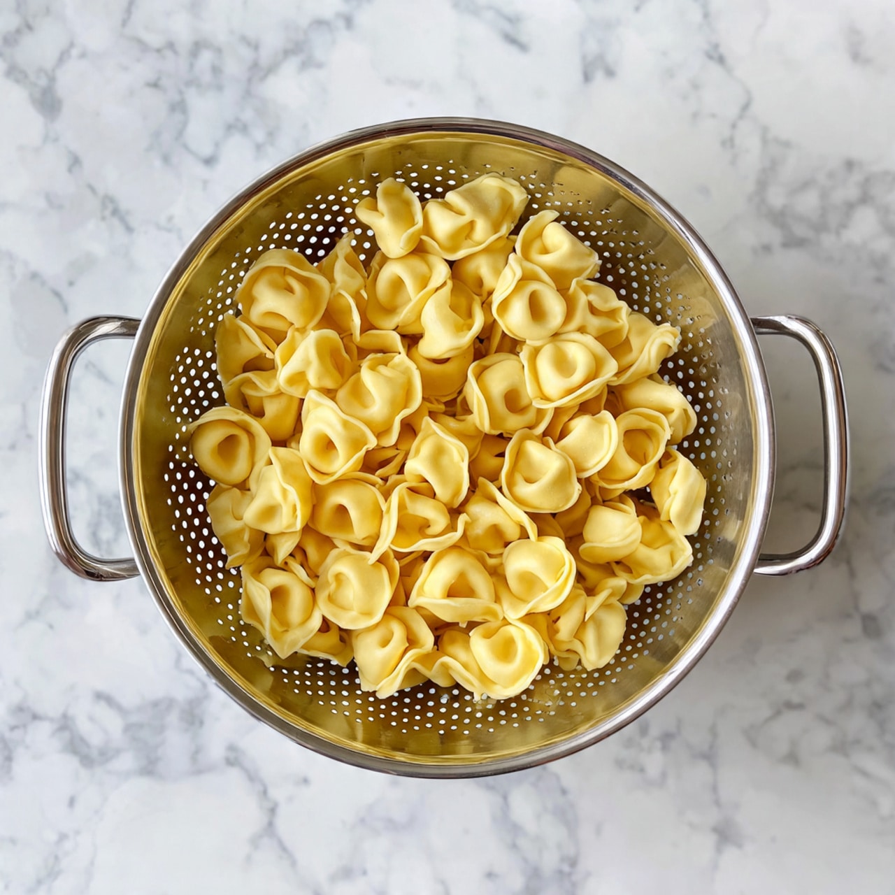 A metal colander filled with one large layer of cooked tortellini pasta, each piece showing smooth, light yellow dough folded into small rings with pinched tops and rounded bottoms, filling the colander evenly. The colander has a shiny, perforated texture with small round holes and is held by two long thin handles extending downward. The colander sits on a white marbled surface, bright and clean. Photo taken with an iphone --ar 4:5 --v 7