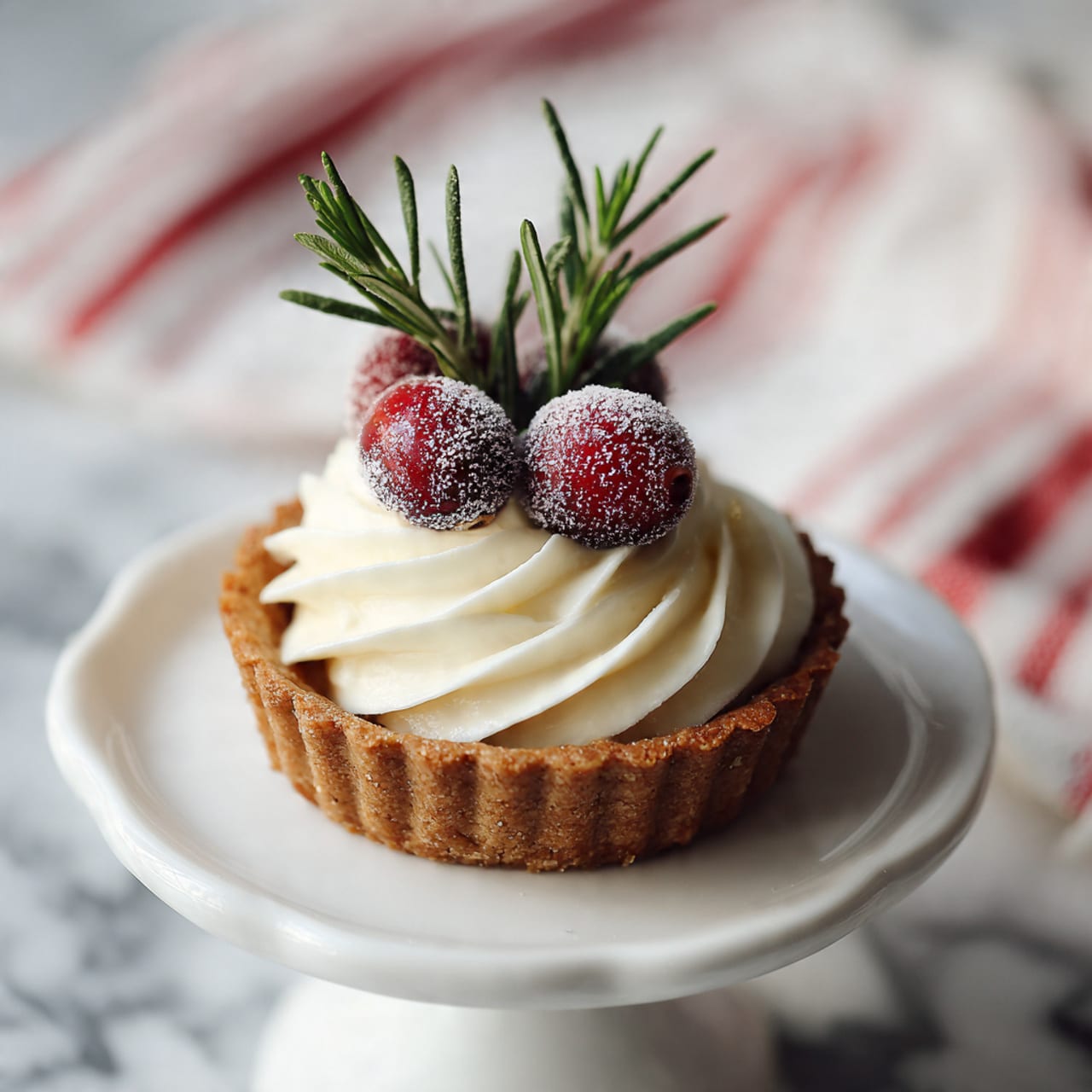 A small round tart with a thick golden brown crust forms the base layer. On top of the crust, there is a thick layer of smooth, white whipped cream, swirled in a circular pattern. Sitting on the whipped cream are three bright red cranberries covered lightly with a sugar coating, giving them a frosty look. Two green rosemary sprigs stand upright behind the cranberries, adding a fresh touch. The tart is placed on a white cake stand with a clean white marbled surface and a red and white striped cloth is blurred in the background. photo taken with an iphone --ar 4:5 --v 7