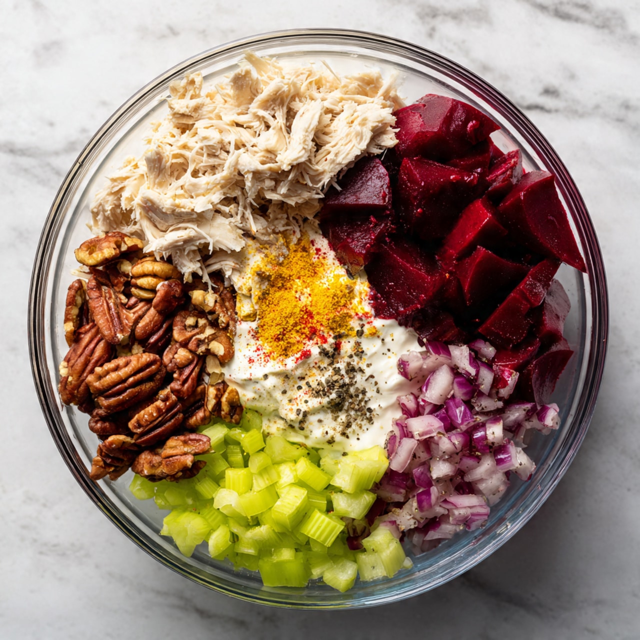 A clear glass bowl on a white marbled surface holds six distinct ingredient groups arranged side by side without mixing. Starting at the top, there is a light beige shredded chicken layer with a soft texture. To the right of the chicken, there are grape halves in a deep red-purple color with a shiny surface. Below the grapes, there is a collection of small, chopped red onions with a mix of purple and white pieces. Moving left, a creamy white layer of yogurt or mayo sits underneath a sprinkle of orange-yellow powder. To the left of that, there is a generous cluster of chopped brown pecans with rough, uneven edges. Finally, to the left, small diced pale green celery pieces topped with black pepper complete the arrangement. Photo taken with an iphone --ar 4:5 --v 7