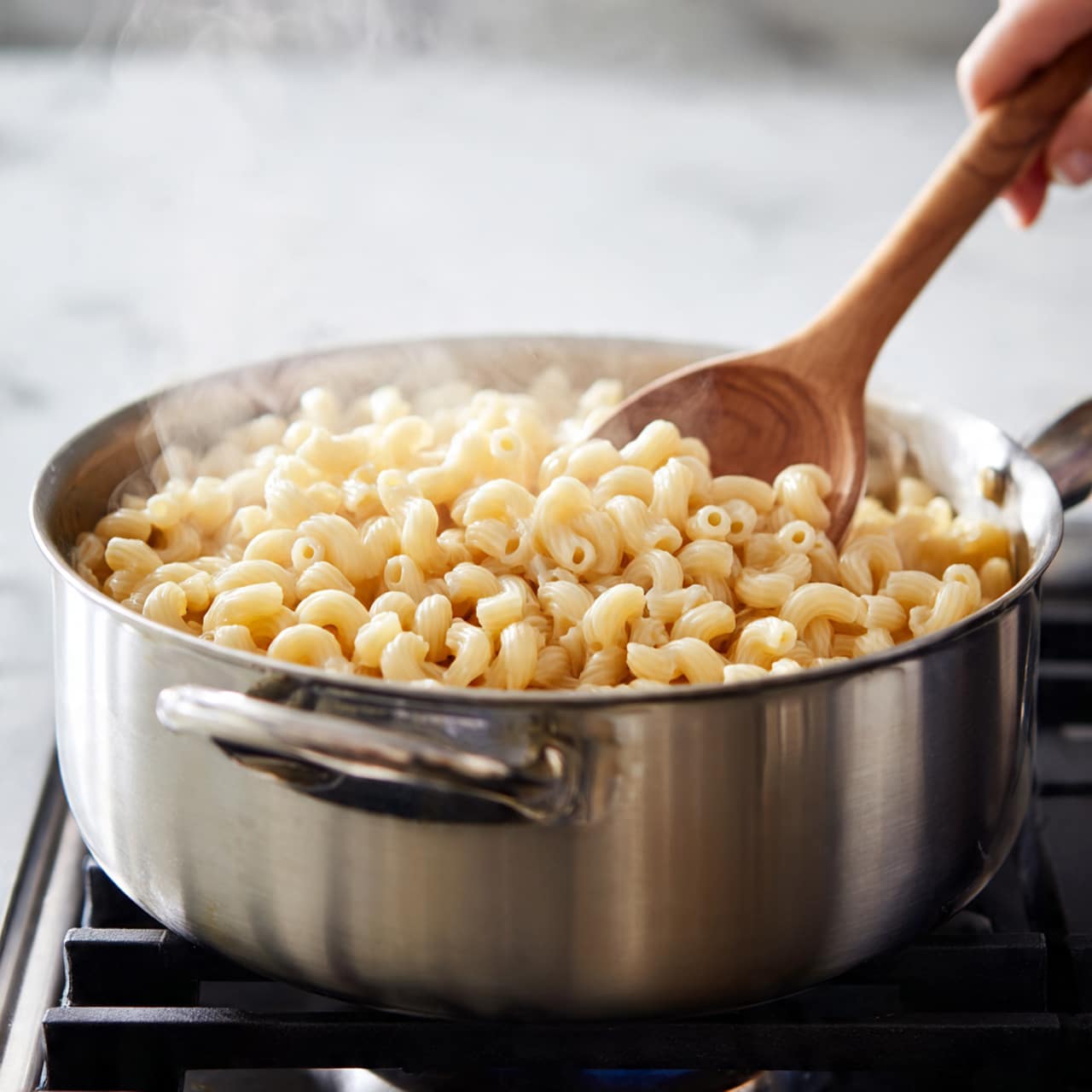 A close-up view of a large silver pot on a stove with black grates, filled with cooked elbow macaroni pasta that is light yellow and soft-looking. A woman's hand holds a wooden spoon stirring the pasta inside the pot. The background surface is white marbled texture. photo taken with an iphone --ar 4:5 --v 7