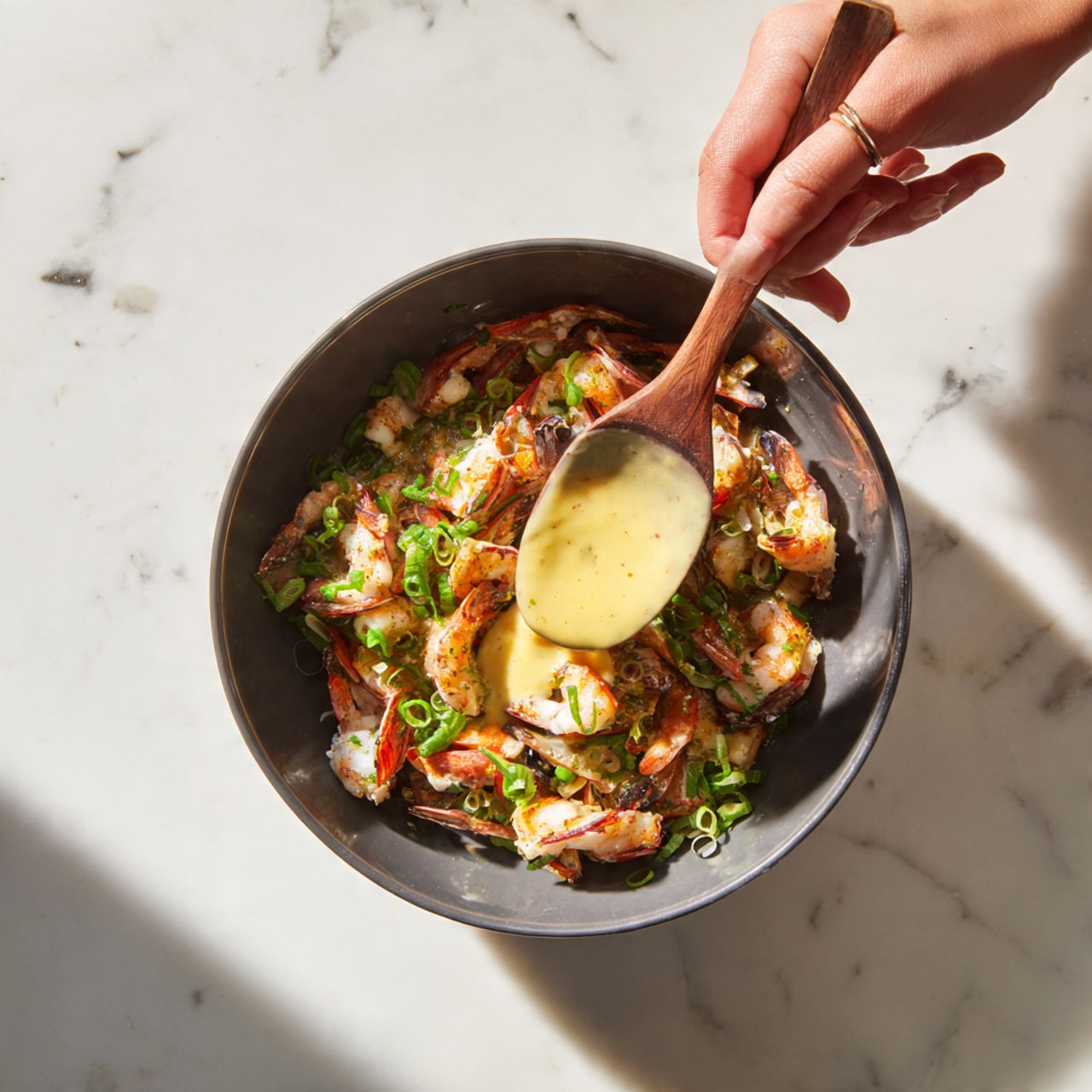 A close-up view of a dark gray bowl filled with small pieces of cooked chicken mixed with chopped green onions and some other small bits of vegetables. A woman's hand holds a wooden spatula inside the bowl, stirring the mixture. From the right side, a pouring container is held by a woman's hand slowly adding a light yellow creamy sauce over the chicken mix. The bowl is placed on a white marbled surface. Photo taken with an iphone --ar 4:5 --v 7