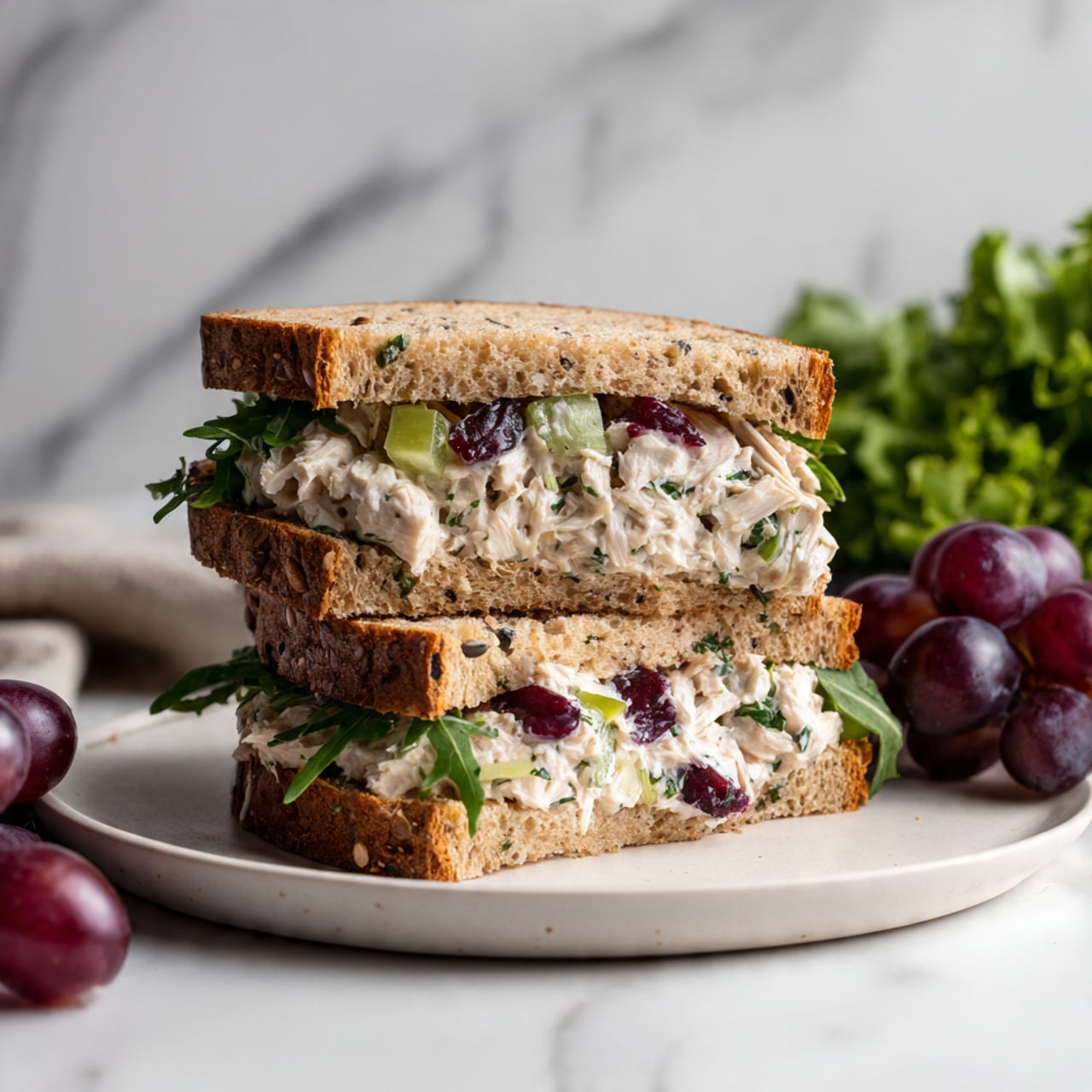 The image shows a sandwich cut in half and stacked with two layers of whole grain bread. Between the bread layers, there is a thick filling made of shredded white chicken mixed with a creamy white dressing, pieces of red grapes, and small bits of green celery. The sandwich is placed on a round white plate with a side of dark purple grapes and a small bunch of green leafy salad. The background is a white marbled surface, and soft light highlights the textures of the bread and the creamy filling. photo taken with an iphone --ar 4:5 --v 7