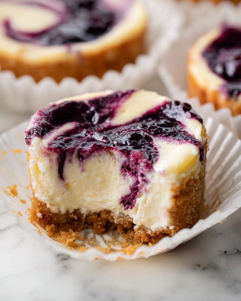The image shows a close-up of a small cheesecake with three visible layers. The bottom layer is a light brown crumbly crust. The middle layer is creamy white cheesecake filling with a smooth texture. Swirled into the top layer is a dark purple blueberry sauce, creating a marbled effect across the surface. A small bite is taken out of the cheesecake, revealing the thick, soft filling and dense crust. The cheesecake sits inside a white paper liner with a crimped edge. The background is a white marbled texture with part of another cheesecake visible in the distance. photo taken with an iphone --ar 4:5 --v 7