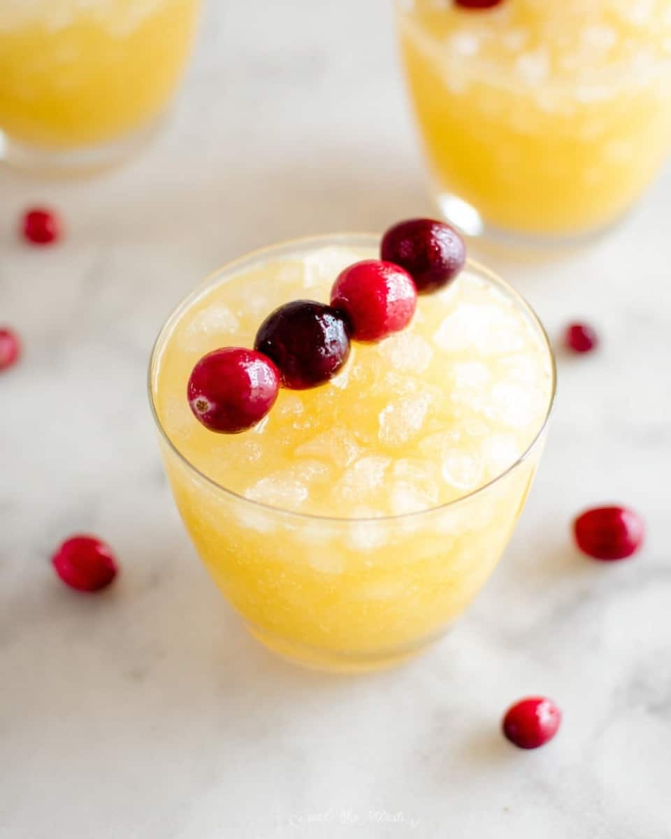 The image shows a clear glass filled with a yellow drink topped with crushed ice. The drink layer is smooth and light yellow, filling about three-quarters of the glass. Three glossy red cranberries and one dark purple berry are placed on top across the rim in a line, creating a colorful contrast. There are more red cranberries scattered on a white marbled surface beneath the glass, with two more glasses of the same drink blurred in the background. The overall scene is bright and clean, with soft natural light highlighting the texture of the ice and the gloss of the berries photo taken with an iphone --ar 4:5 --v 7