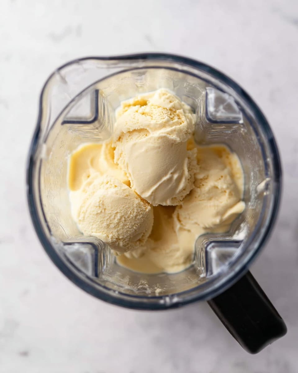 The image shows a top view of a clear blender jar with several scoops of creamy light yellow ice cream inside. The blender jar has distinct sharp edges and a black handle on the right side. The ice cream texture looks smooth with some rounded and soft uneven parts. The surface beneath is white with a subtle marbled pattern. photo taken with an iphone --ar 4:5 --v 7