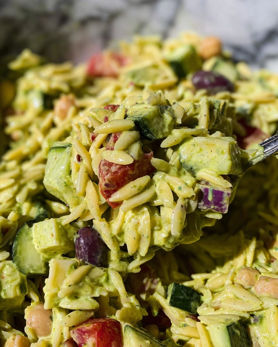 This image shows a close-up of a creamy orzo salad with visible layers of yellow orzo pasta mixed with a thick green-yellow dressing. Cubes of fresh green cucumber and pieces of red tomato are spread evenly throughout the salad, along with some purple olives and chickpeas adding color contrast. The salad appears moist, with a glossy finish on all ingredients. The mixture is captured being scooped, showing a dense, textured, and creamy composition. The background features a white marbled texture. photo taken with an iphone --ar 4:5 --v 7
