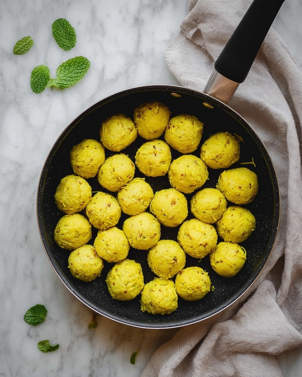 A white plate holds a bed of fluffy white rice with long grains, arranged to cover the base of the plate evenly. On top, there are four round yellow curry meatballs, coated in a thick sauce with specks of green herbs scattered over them. Around the plate, a few fresh green mint leaves are placed on the white marbled texture surface. A light gray cloth rests to the right side near the plate. The dish looks warm and inviting, with a mix of soft textures and vibrant colors. photo taken with an iphone --ar 4:5 --v 7