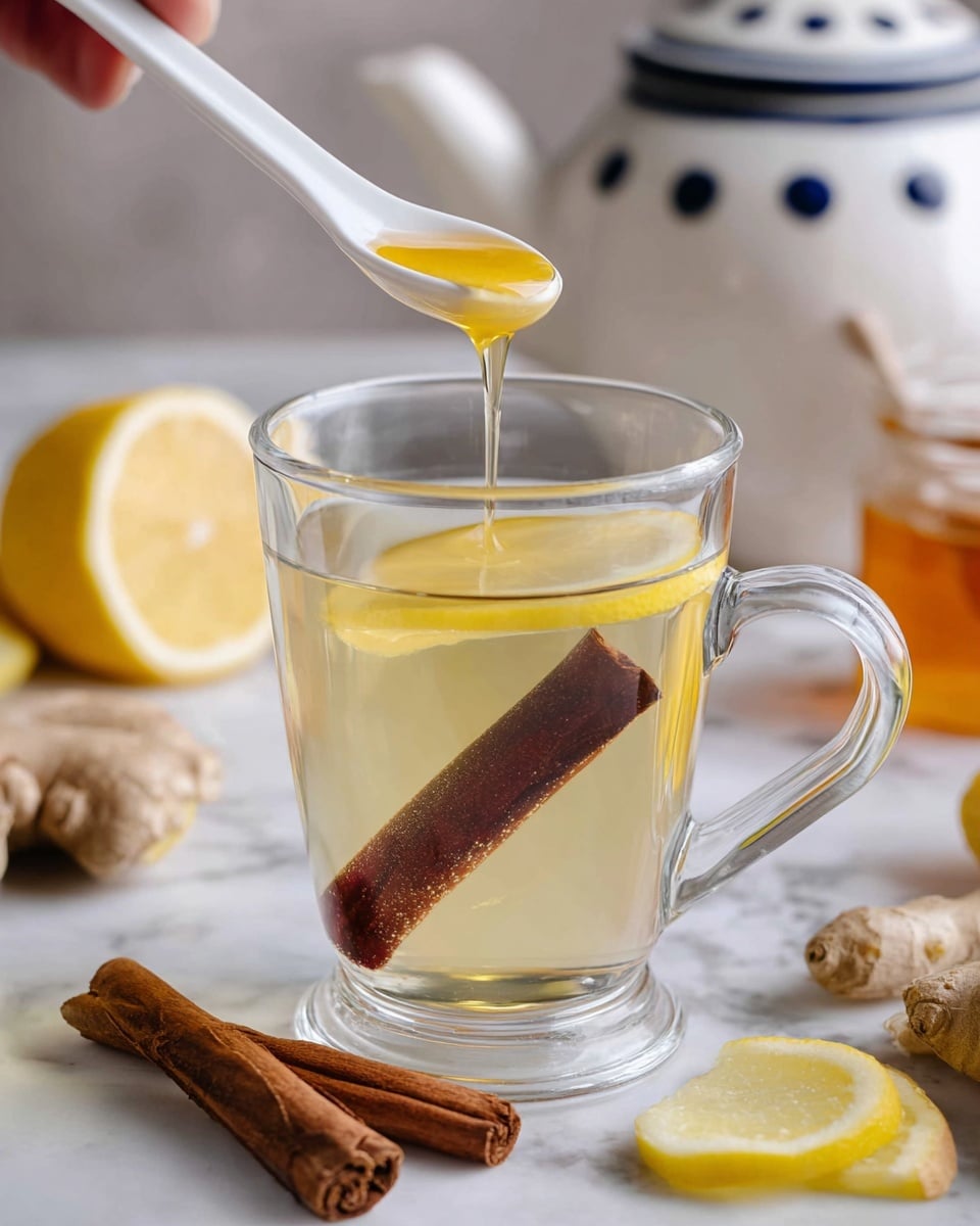 A clear glass cup filled with light yellow tea contains a large dark brown cinnamon stick standing upright inside and a thin bright yellow lemon slice floating near the top. A white ceramic spoon held by a woman's hand drizzles golden honey into the tea from above. The glass cup sits on a white marbled surface, with two cinnamon sticks lying nearby, along with pieces of fresh ginger and a halved lemon. In the background, out of focus, is a white ceramic teapot with blue dots and a small jar with golden honey visible near the bottom right corner. photo taken with an iphone --ar 4:5 --v 7
