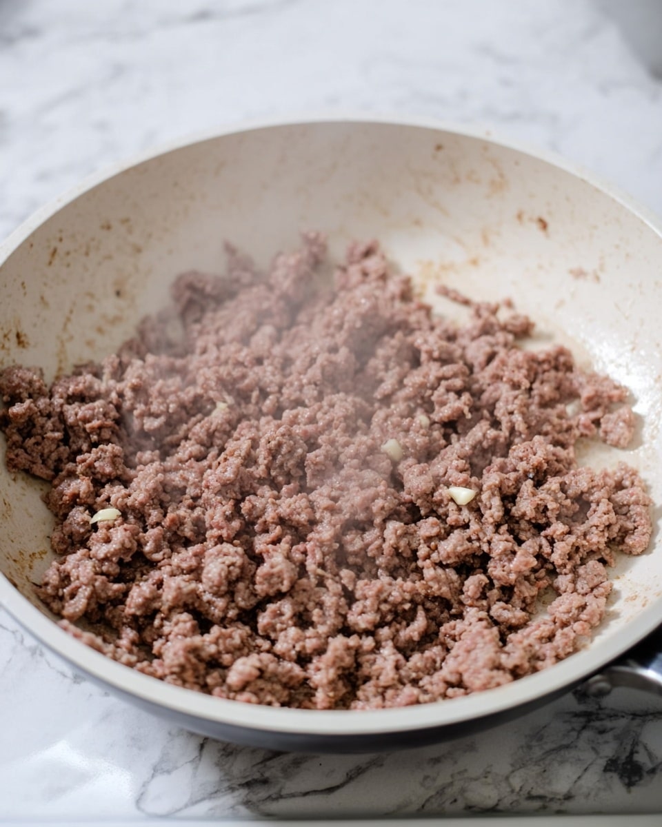 Inside a white frying pan, there is a layer of cooked ground beef with a brown and slightly pink color, showing a crumbly texture. Small pieces of minced garlic are mixed in with the beef, visible as off-white bits. Steam rises gently from the pan, which sits on a stovetop. The background is a white marbled texture. photo taken with an iphone --ar 4:5 --v 7