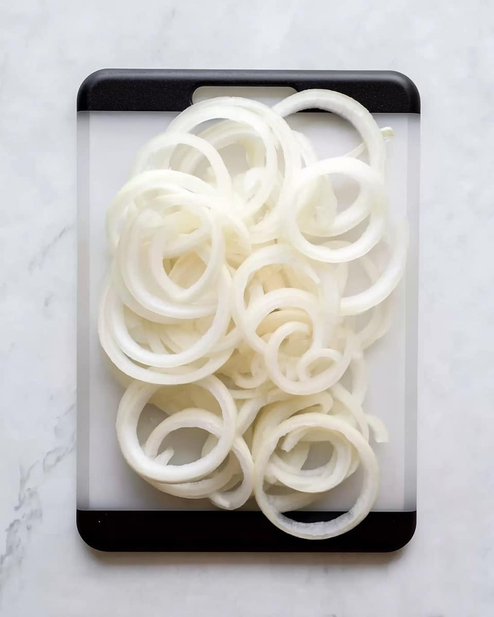 The image shows a group of thin, smooth onion rings arranged loosely on a clear rectangular cutting board with black corners. The onion rings vary in size, with some large rings at the edges and smaller, more tightly curled rings towards the center. The cutting board is placed on a white marbled surface, giving a clean and bright look to the setting. The onion rings have a pale white to light translucent color with soft textures, and their round shapes create a layered pattern across the board. Photo taken with an iphone --ar 4:5 --v 7