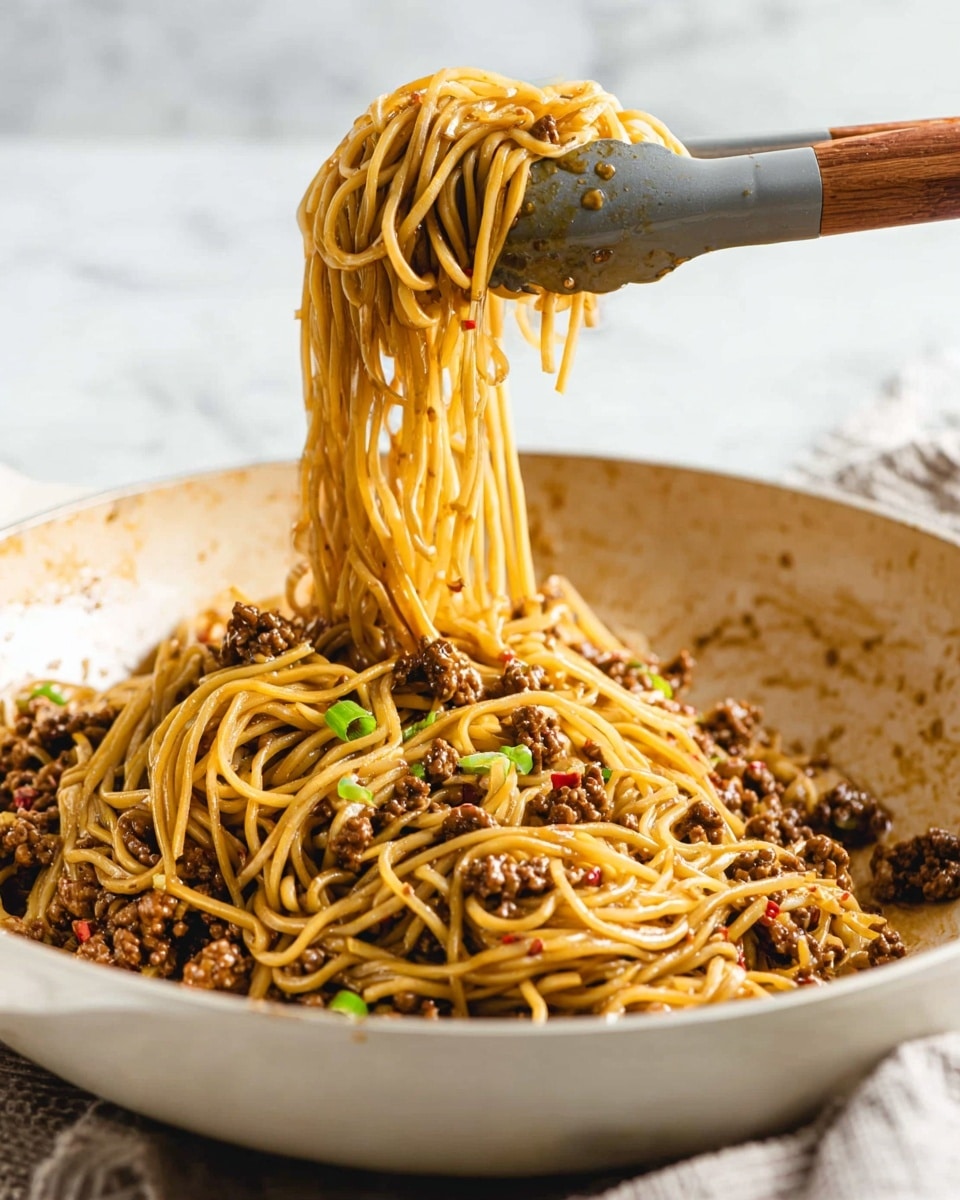 A white pan holds a large serving of thin, yellowish-brown noodles mixed with small pieces of cooked ground meat that are dark brown. The noodles and meat are coated evenly in a glossy brown sauce with small red chili flakes and bits of green onions scattered throughout. A gray tong with a wooden handle lifts a bundle of the noodles above the pan, showing their long and smooth texture as they drip slightly. The background features a soft white marbled surface, enhancing the warm tones of the dish. Photo taken with an iphone --ar 4:5 --v 7