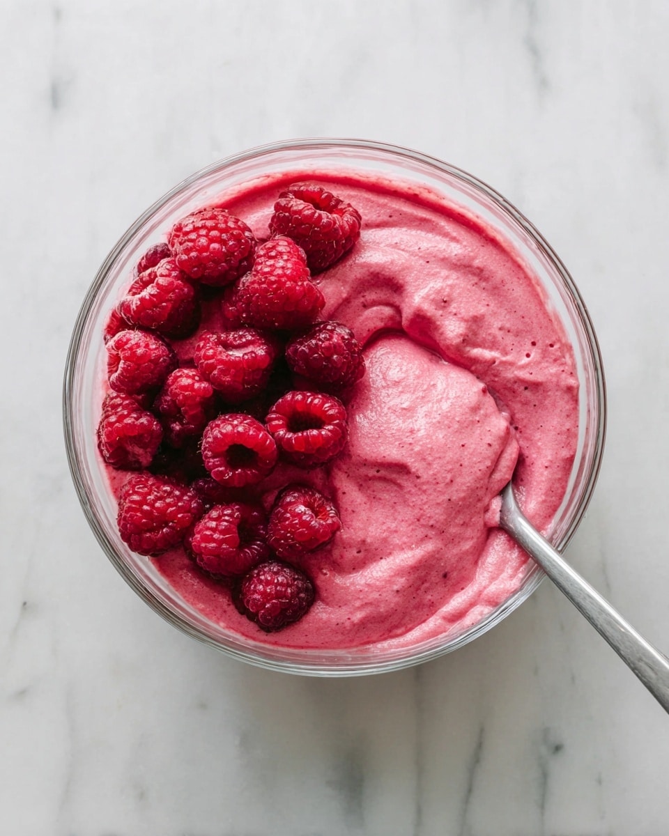 A clear glass bowl filled with a thick, creamy bright pink mixture that has a smooth, slightly textured surface. On top, there is a cluster of fresh whole raspberries with a deep red color positioned mostly on the left side of the bowl. A metal spoon resting inside the bowl is partially covered with the bright pink mixture and leans on the right side. The bowl is placed on a white marbled surface with subtle gray veins. Photo taken with an iphone --ar 4:5 --v 7