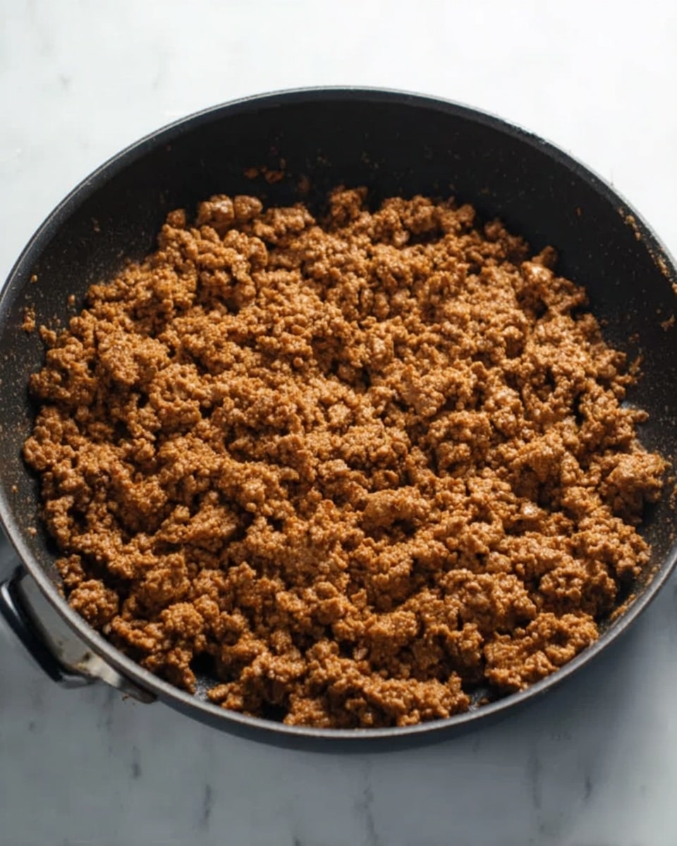 A black pan filled with cooked ground meat, the meat is brown and crumbly with a slightly rough texture. The pan has a simple design with small handles on the sides. The background is a white marbled surface, clean and bright, making the pan and meat stand out clearly. There are no extra ingredients or garnishes visible, just the cooked meat spread evenly across the pan. photo taken with an iphone --ar 4:5 --v 7