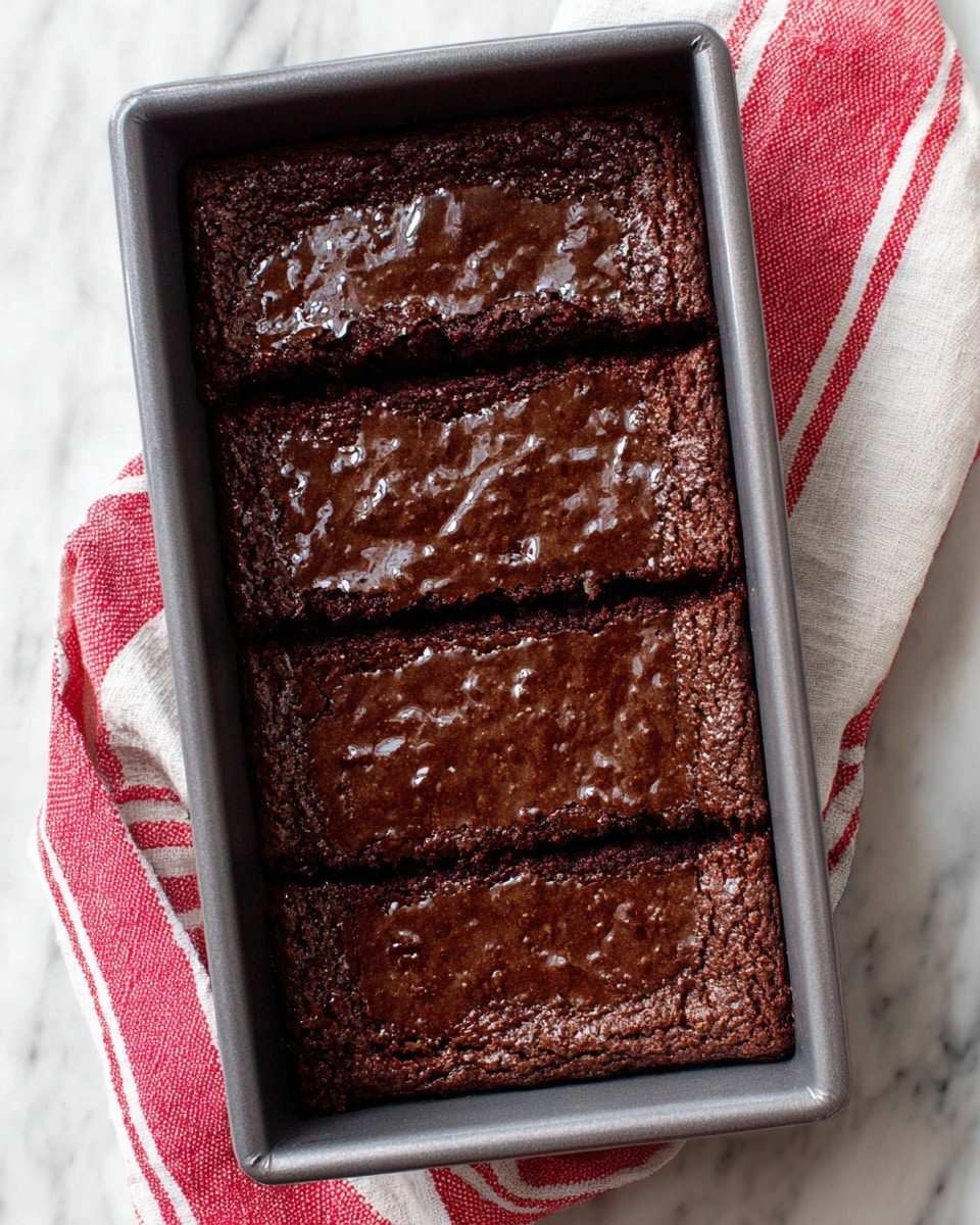 The image shows a dark brown baked dessert with a shiny, slightly cracked top layer in a gray rectangular baking pan. The dessert has three rectangular sections, divided by two straight cuts across its width. The texture appears moist and firm with a glossy surface that catches the light. The pan is placed on a white marbled surface with a red and white striped cloth partially visible underneath. Photo taken with an iphone --ar 4:5 --v 7