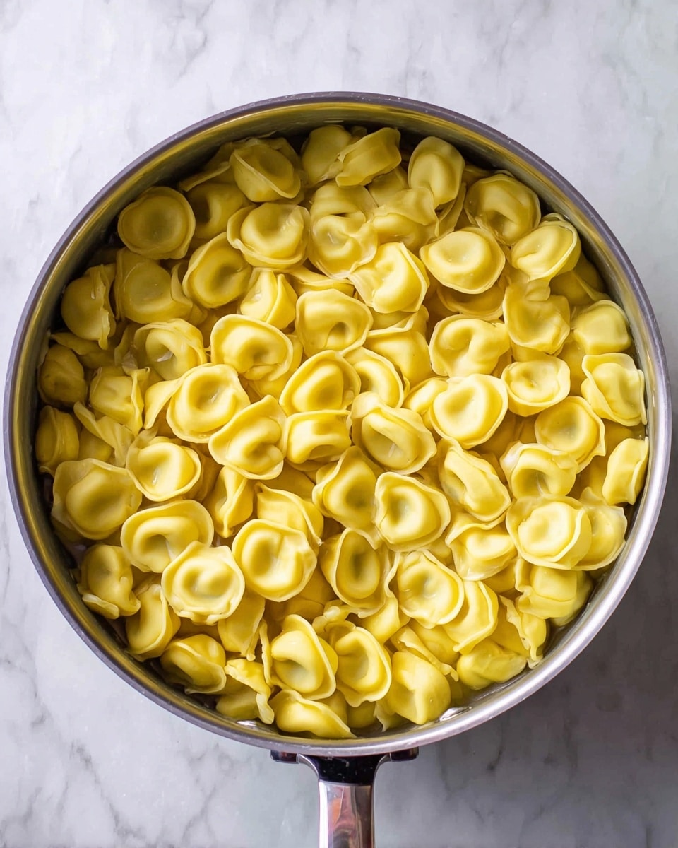 A close-up top view of a large silver pot filled with one layer of yellow tortellini pasta, each piece having a smooth and slightly shiny texture, sitting in water. The pot handle is seen at the right side on a white marbled surface. Photo taken with an iphone --ar 4:5 --v 7