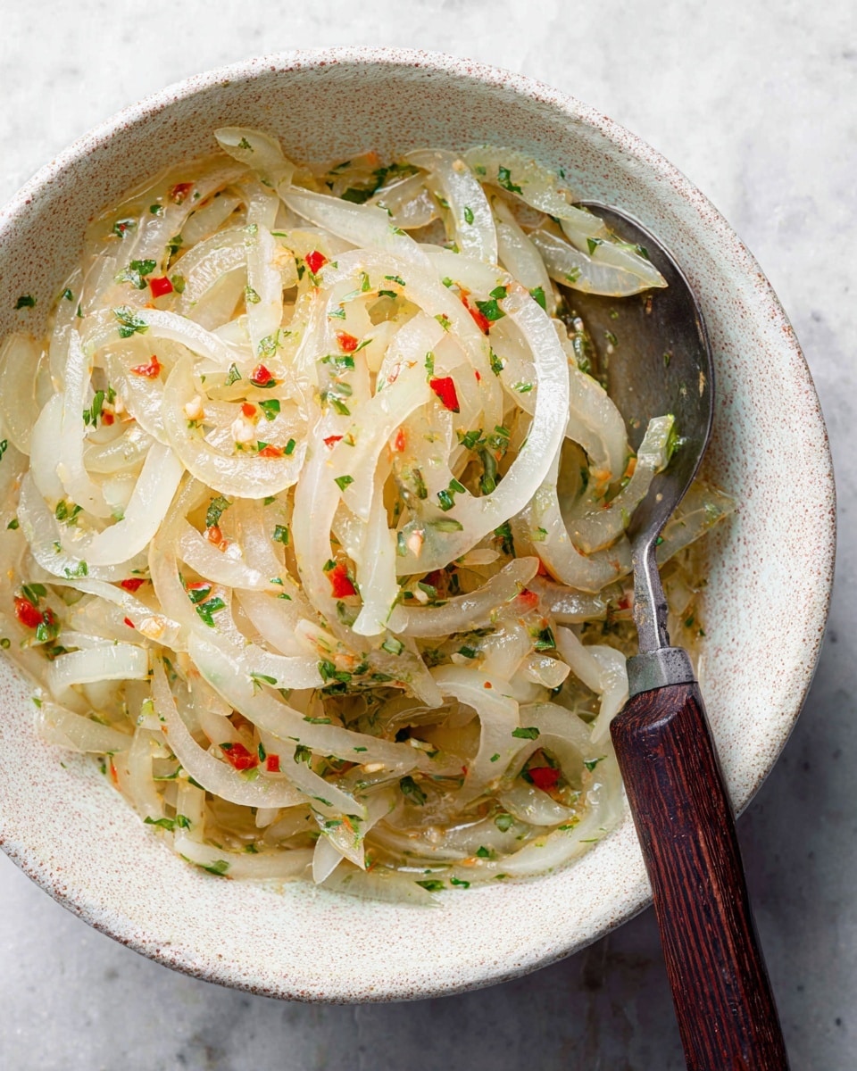 A white speckled bowl filled with thin, translucent slices of cooked onions mixed with small pieces of red chili and green herbs evenly scattered throughout. The onions have a soft shine from light oil or dressing, creating a slightly wet texture. A spoon with a dark wooden handle rests inside the bowl on the right side. The bowl sits on a white marbled surface. photo taken with an iphone --ar 4:5 --v 7