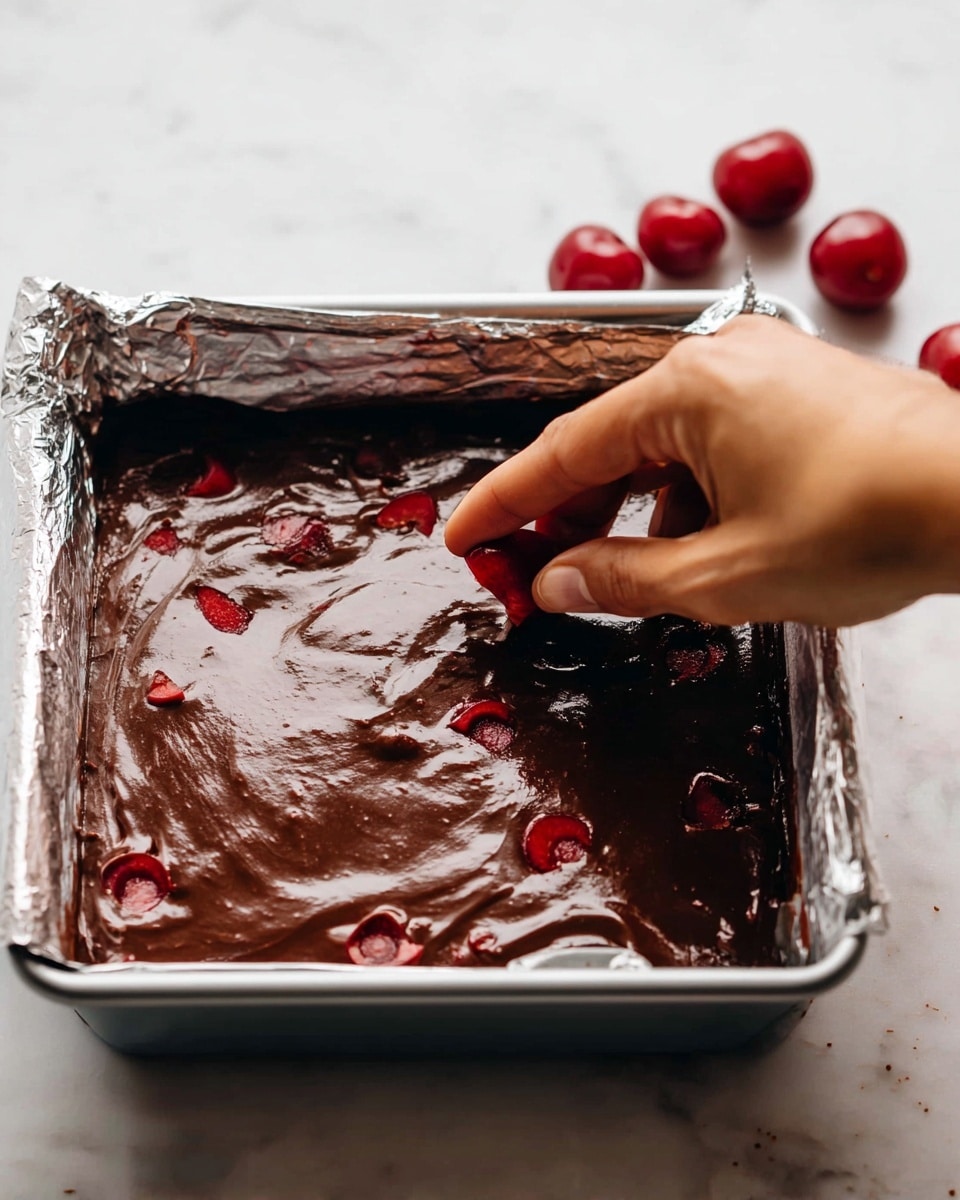 A close-up view of a shiny dark brown chocolate batter spread evenly inside a silver square baking pan lined with foil, with visible small red cherry slices mixed into the batter layer. A woman's hand is gently pressing one cherry slice into the thick batter in the top right corner of the pan. In the background, there are a few whole red cherries scattered on a white marbled surface. The overall look is moist and glossy, highlighting the texture of the batter and fruit pieces. Photo taken with an iphone --ar 4:5 --v 7