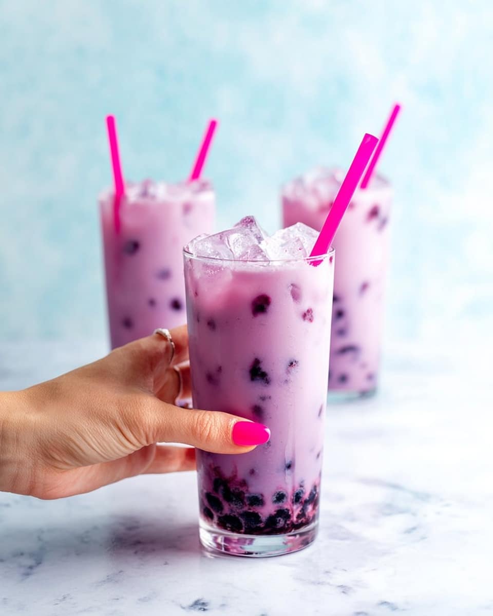 The image shows three clear tall glasses filled with a light purple smoothie that has dark purple and black berry pieces mixed inside. Each glass is layered with ice cubes near the top, making the drink look cold and fresh. Bright pink bendy straws stick out from the glasses, adding a pop of color. A woman’s hand with bright pink nail polish holds the front glass, which is in sharp focus while the two behind it are slightly blurred. The glasses are placed on a white marbled surface with a soft, light blue and white background. photo taken with an iphone --ar 4:5 --v 7