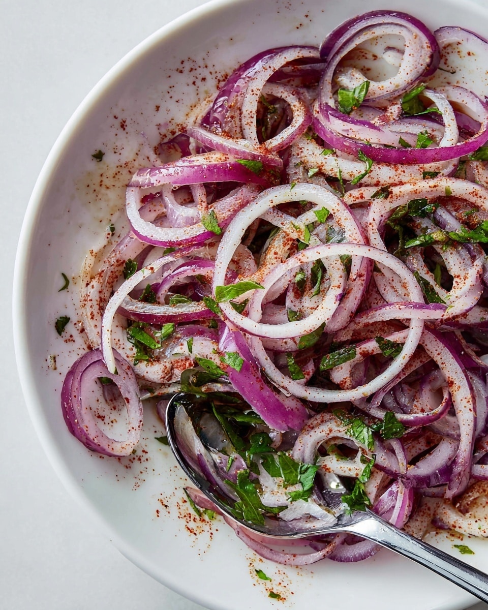 A close-up view of a white plate filled with thinly sliced rings of purple and light pink onions mixed with small pieces of bright green herbs. The onion rings are coated lightly with a reddish spice powder, scattered evenly throughout. A shiny silver spoon rests on the right side, partly under the onion slices, reflecting some of the colors. The plate sits on a white marbled surface, adding a clean and fresh look. Photo taken with an iphone --ar 4:5 --v 7
