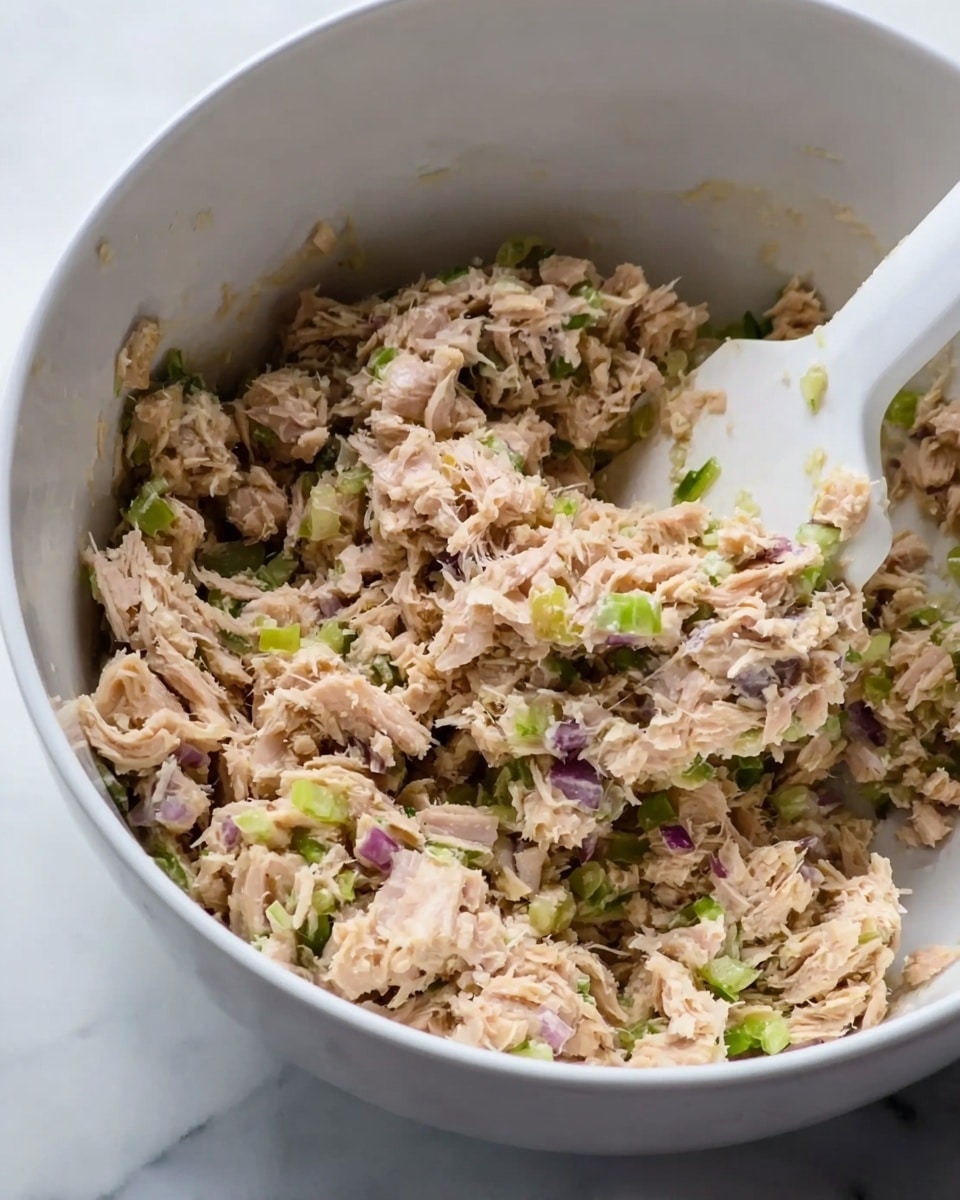 The image shows a close-up view of a white bowl filled with a mixture of shredded light brown tuna flakes, small bits of green celery, and tiny pieces of purple onion, all combined together with a white spatula partially visible on the right side stirring the ingredients. The texture appears soft and moist in the evenly mixed layers, set against the inside of the white bowl with a white marbled surface in the background. Photo taken with an iphone --ar 4:5 --v 7