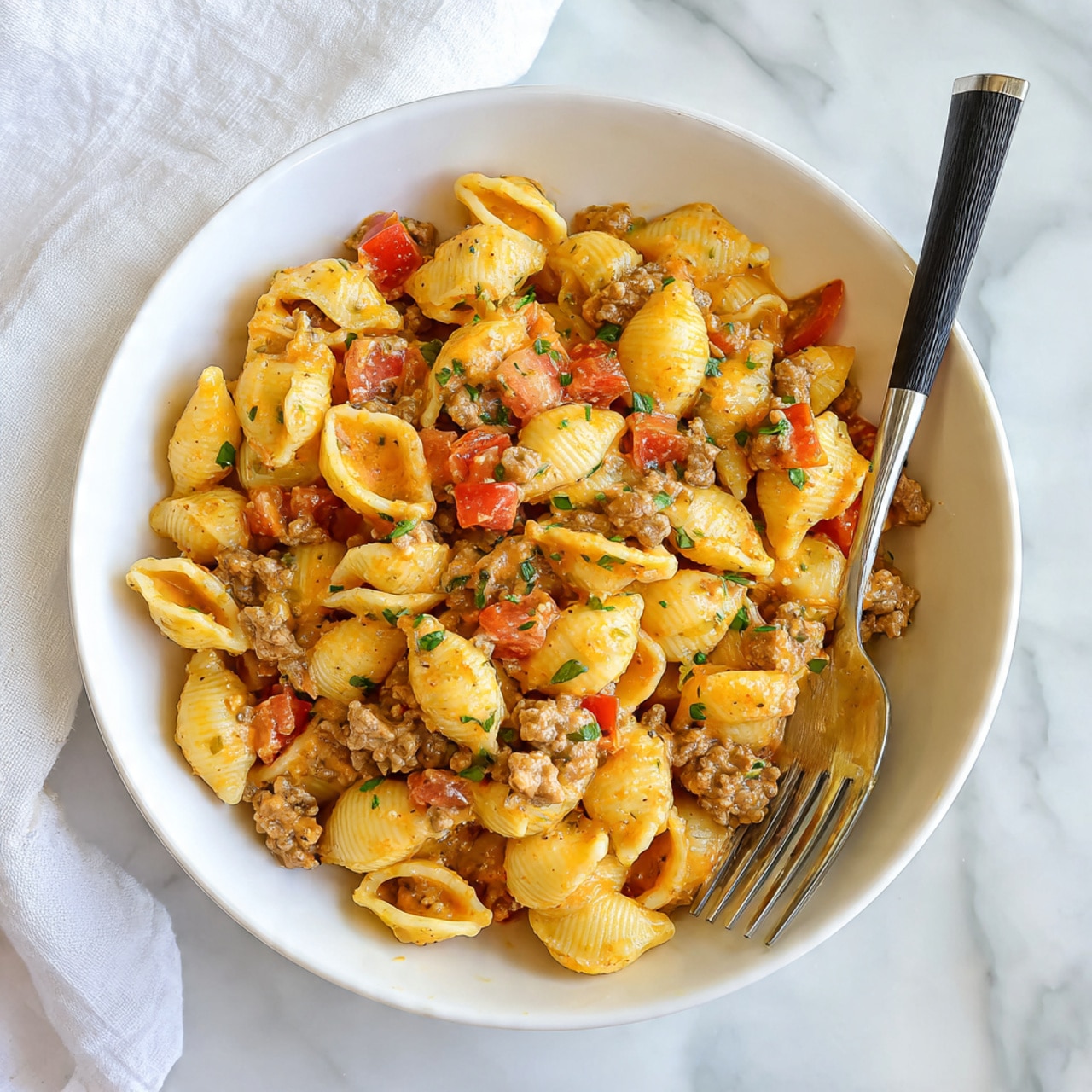 A white bowl filled with about two layers of yellow shell pasta mixed with a creamy orange sauce, ground meat, and chunks of red tomato, all garnished with small green herb pieces. A metal fork with a black handle rests inside the bowl on the right side, and the bowl is placed on a white marbled surface with a white cloth in the background. photo taken with an iphone --ar 4:5 --v 7