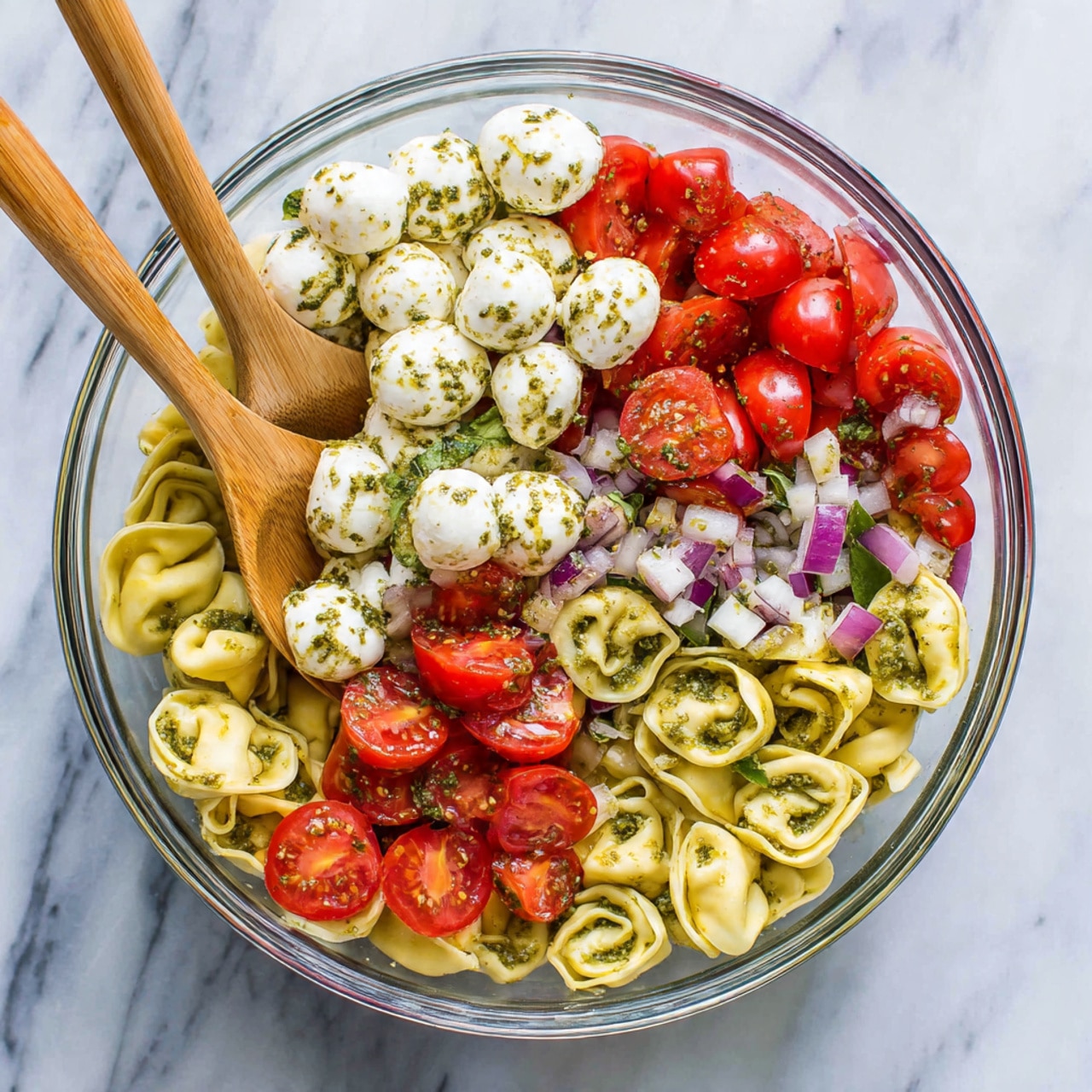 The image shows a clear glass bowl filled with three visible layers of food. The bottom layer has light yellow tortellini pasta with a bit of green pesto sauce on top. The middle layer contains bright red cherry tomatoes, some whole and some sliced, scattered evenly. The top layer is made up of small white mozzarella balls mixed with green basil leaves and small pieces of purple onion, all coated lightly with green specks of dried herbs or pesto. Two wooden spoons are placed inside the bowl, resting on the tortellini. The bowl sits on a white marbled surface. Photo taken with an iphone --ar 4:5 --v 7