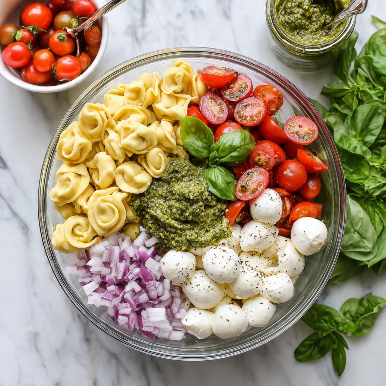 The image shows a clear glass bowl placed on a white marbled surface, filled with different fresh ingredients arranged in separate sections. There are pale yellow tortellini pasta on the left side of the bowl. On top of the pasta, a dollop of green pesto sauce sits in the center left. To the right of the pesto, bright red halved cherry tomatoes fill the right side of the bowl. In the lower right part of the bowl, there are small white mozzarella balls sprinkled with black pepper. In the lower left area, finely chopped purple onions add color. A fresh green basil leaf rests on the onions close to the mozzarella. Behind the bowl, there is a small white bowl with more halved cherry tomatoes and a jar of green pesto with a spoon in it. On the upper right side of the image, bunches of fresh basil leaves rest on the surface. Photo taken with an iphone --ar 4:5 --v 7